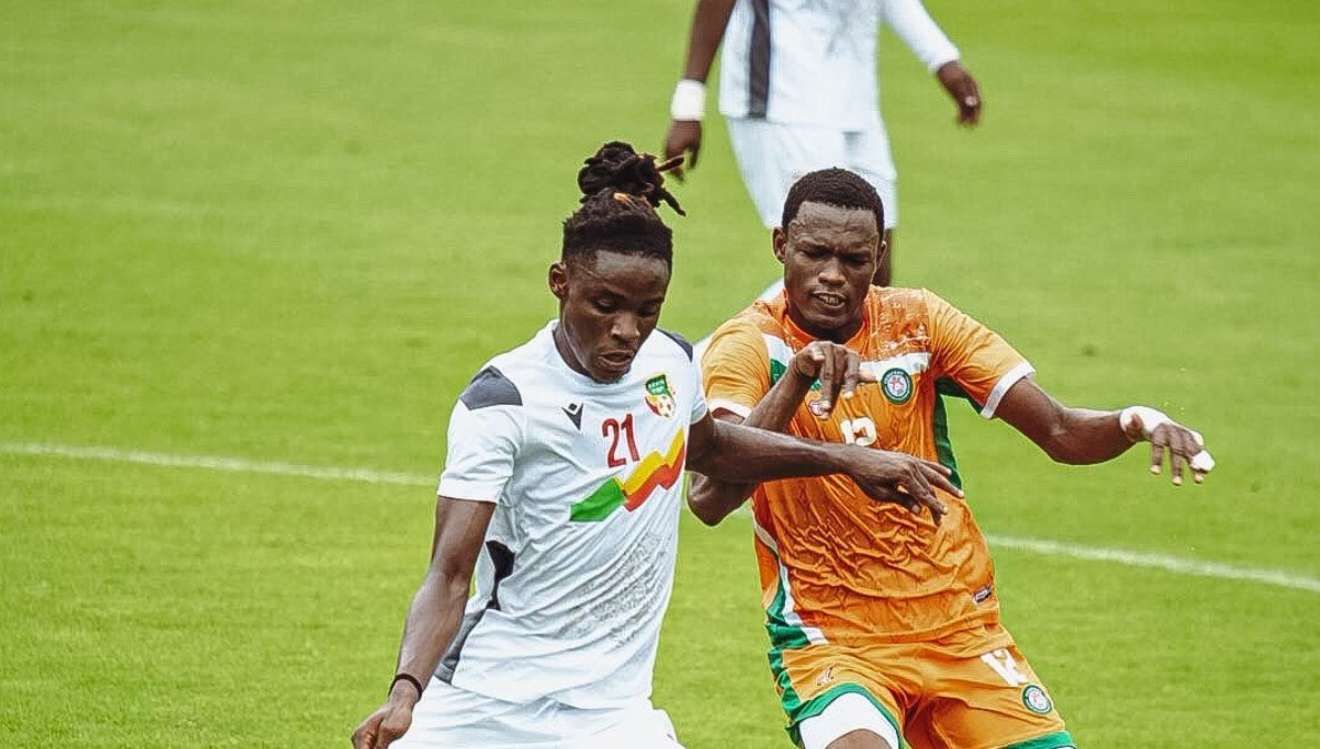 Two soccer players compete for the ball on a green field; one in white kit with the number 21 and the other in orange kit intertwined in a challenge. Two soccer players compete for the ball on a green field; one in white kit with the number 21 and the other in orange kit intertwined in a challenge.