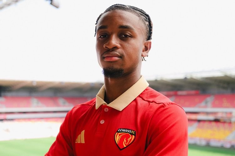 Portrait of a soccer player wearing a red Le Mans FC jersey with a beige collar at a stadium field behind him, looking at the camera.