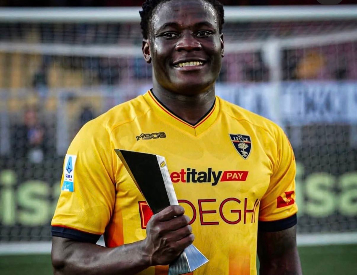 Smiling Black male soccer player in a yellow jersey holding a trophy on a soccer field.