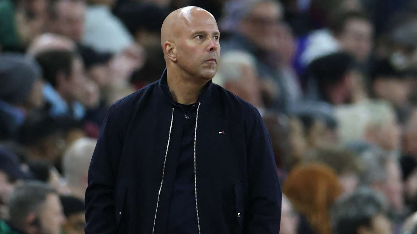 Bald man in a dark jacket stands in a crowded arena, looking to his left.