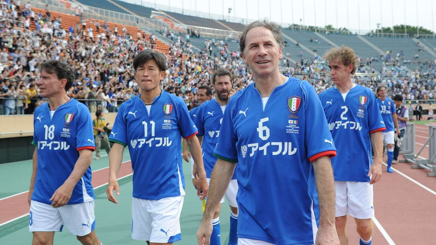 Group of Italian soccer players in blue jerseys walking along a track after a game, with a cheering stadium crowd behind them. Group of Italian soccer players in blue jerseys walking along a track after a game, with a cheering stadium crowd behind them.