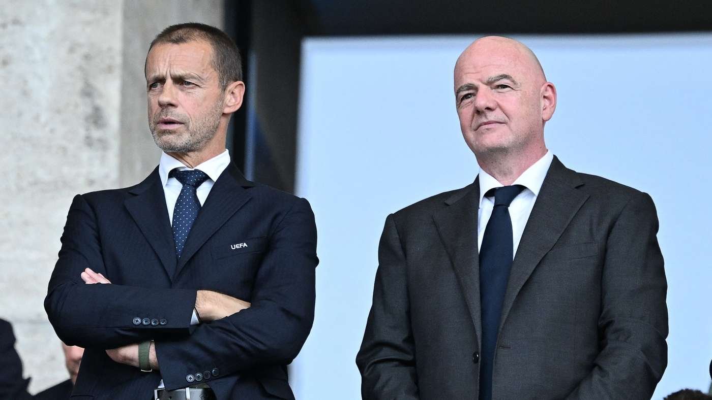 Two men in dark suits stand outdoors, one with arms crossed and the other looking ahead, a UEFA logo visible on the left man's jacket Two men in dark suits stand outdoors, one with arms crossed and the other looking ahead, a UEFA logo visible on the left man's jacket