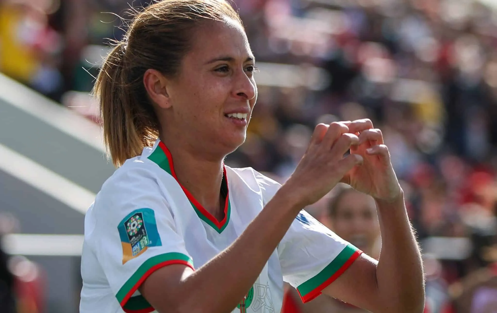 Female athlete in a white jersey with green and red trim claps hands in celebration at a sports event, blurred crowd in background Female athlete in a white jersey with green and red trim claps hands in celebration at a sports event, blurred crowd in background