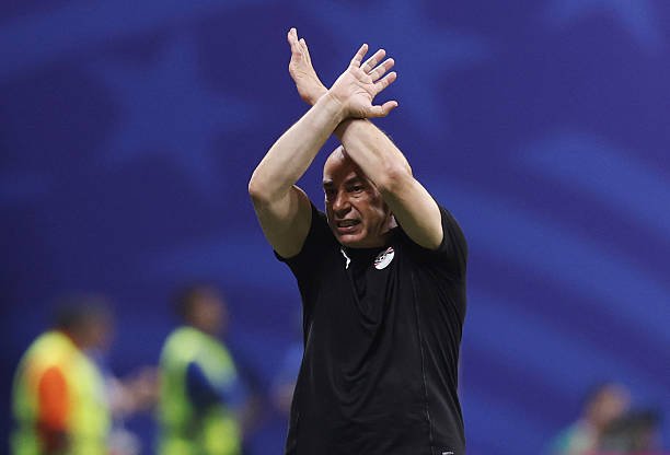 Man in a black shirt crosses his arms above his head during a sports event against a blue backdrop