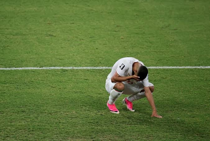 Soccer player in white kit kneeling on grass with head bowed and hand to face, showing disappointment or fatigue. Soccer player in white kit kneeling on grass with head bowed and hand to face, showing disappointment or fatigue.