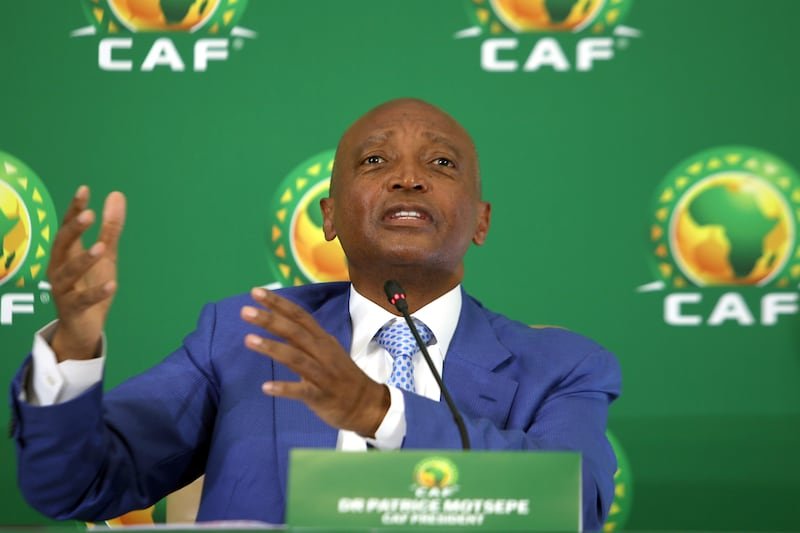 Man in a blue suit speaks at a CAF press conference, gesturing with his hands in front of a green backdrop with CAF logos and a microphone nearby.