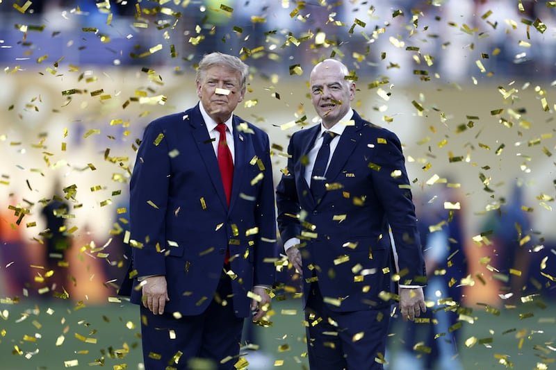 Two men in dark suits pose on a grass field amid falling gold confetti at a celebration.