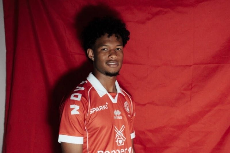 Portrait of a young man in a red soccer jersey with a white collar and cuffs, standing in front of a red backdrop.