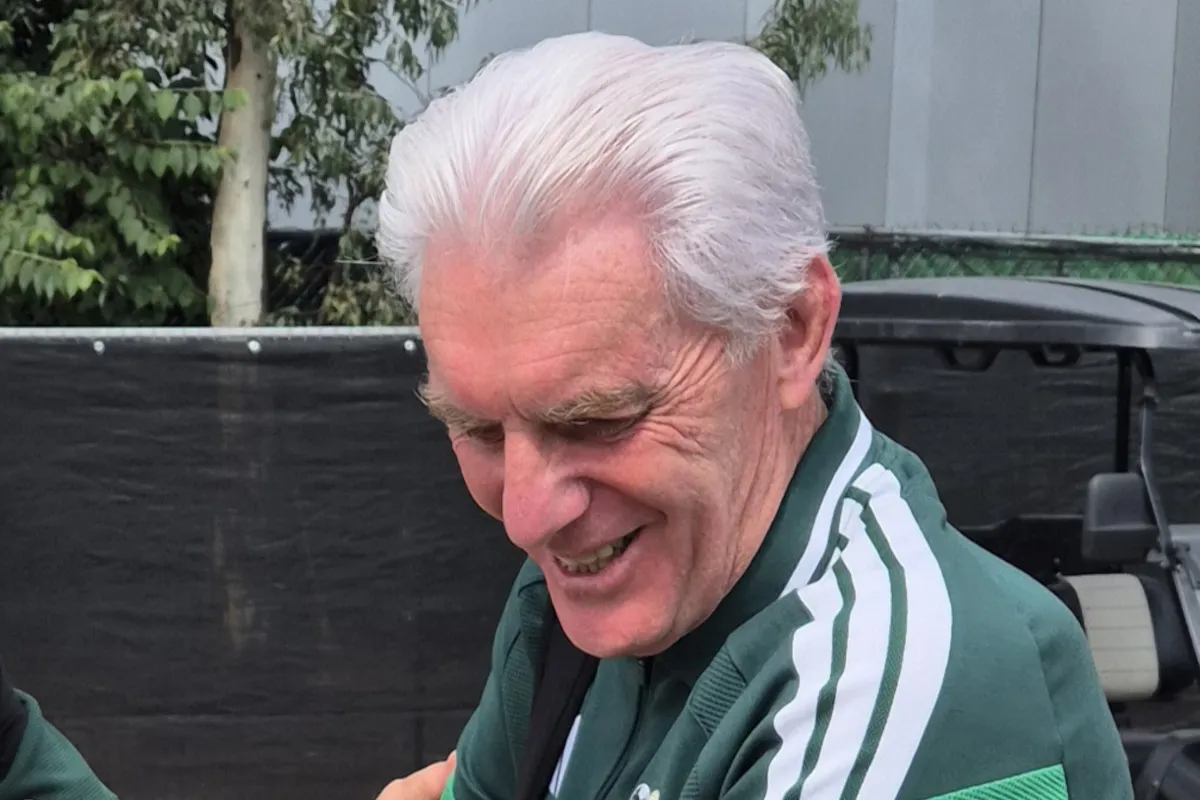Smiling older man with white hair in a green track jacket with white stripes, outdoors near a golf cart area.
