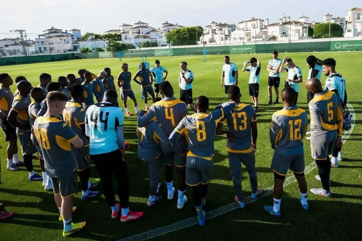 Soccer players in gray and yellow uniforms form a circle huddle on a sunny training field as coaches stand nearby.