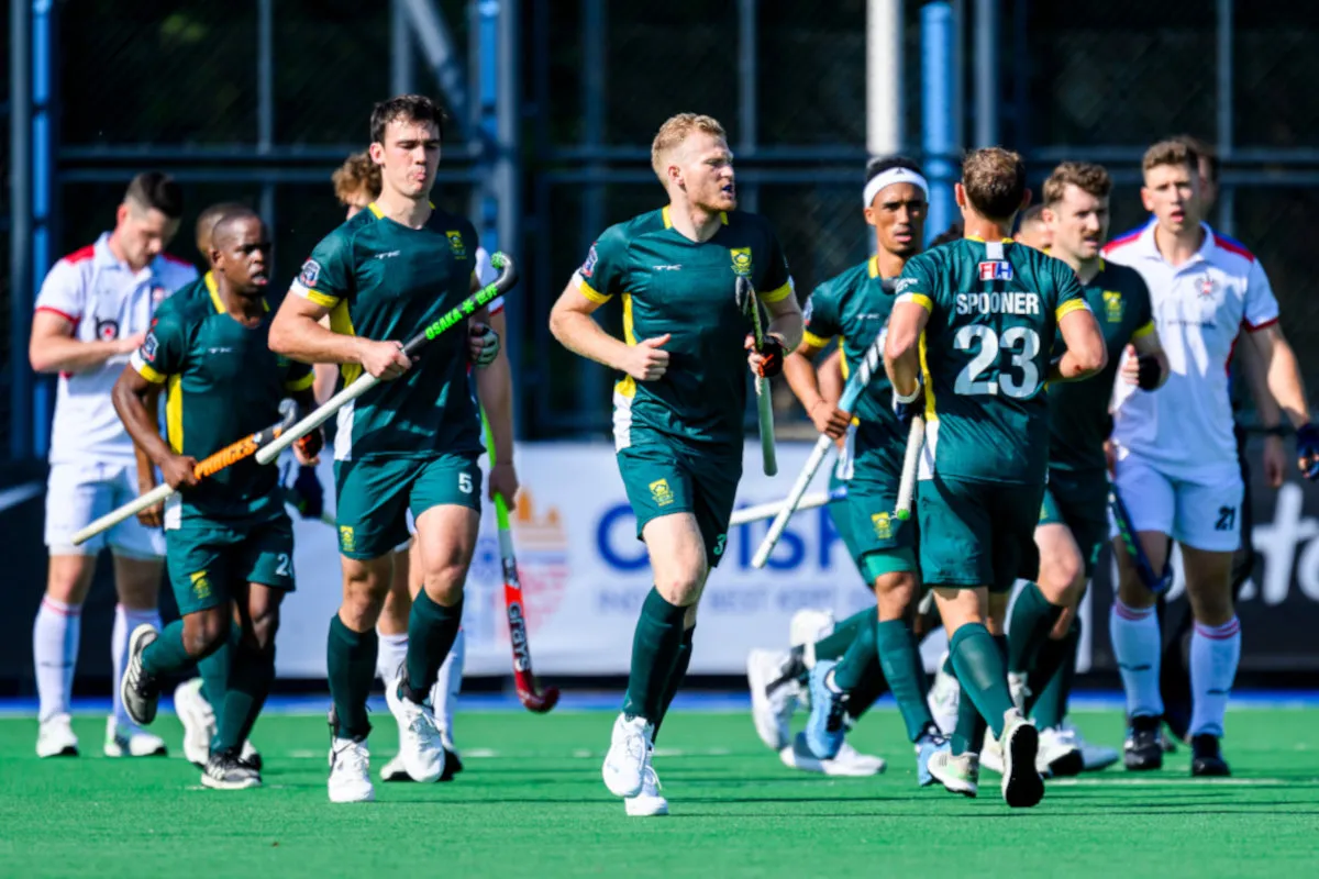 Group of male field hockey players in green uniforms run together on a turf pitch, sticks in hand after a match.