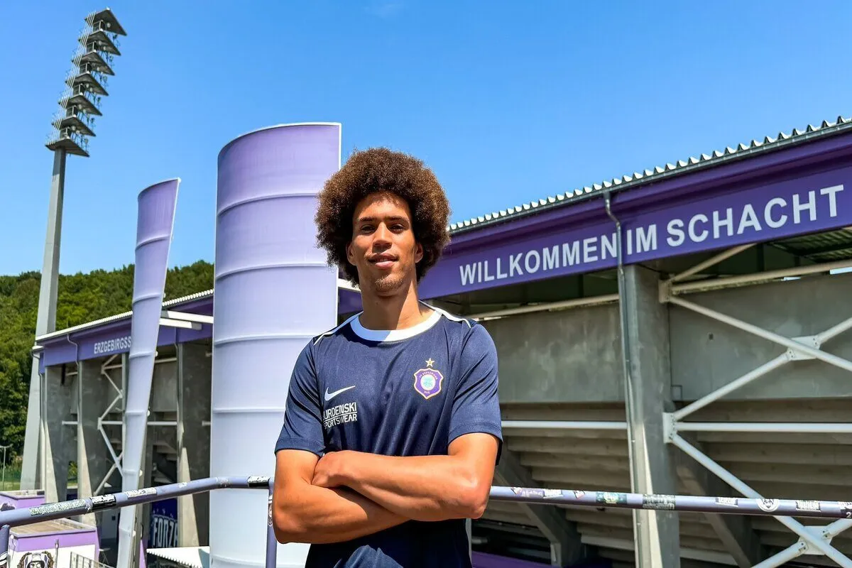 Young man with curly hair stands with arms crossed in front of a purple stadium entrance and a banner reading 'WILLKOMMEN IM SCHACHT'.