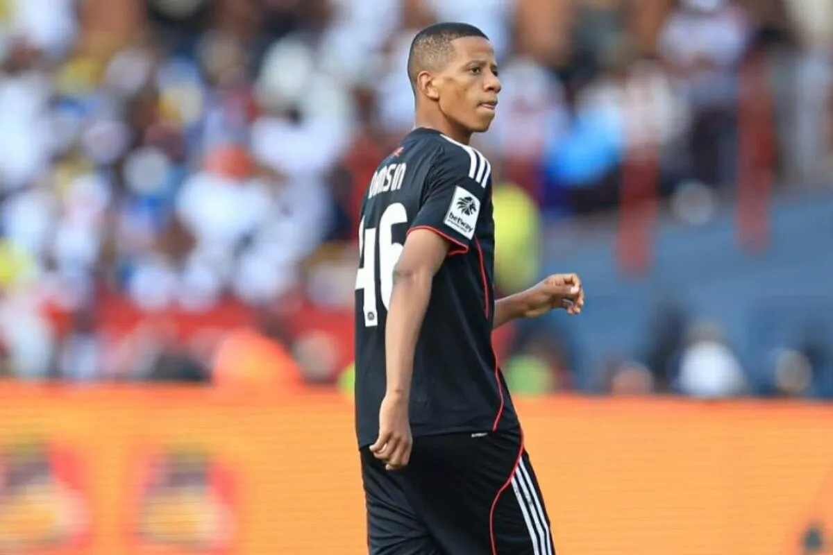 Soccer player in a black kit with white stripes and the number 46, standing on the field as blurred fans watch.
