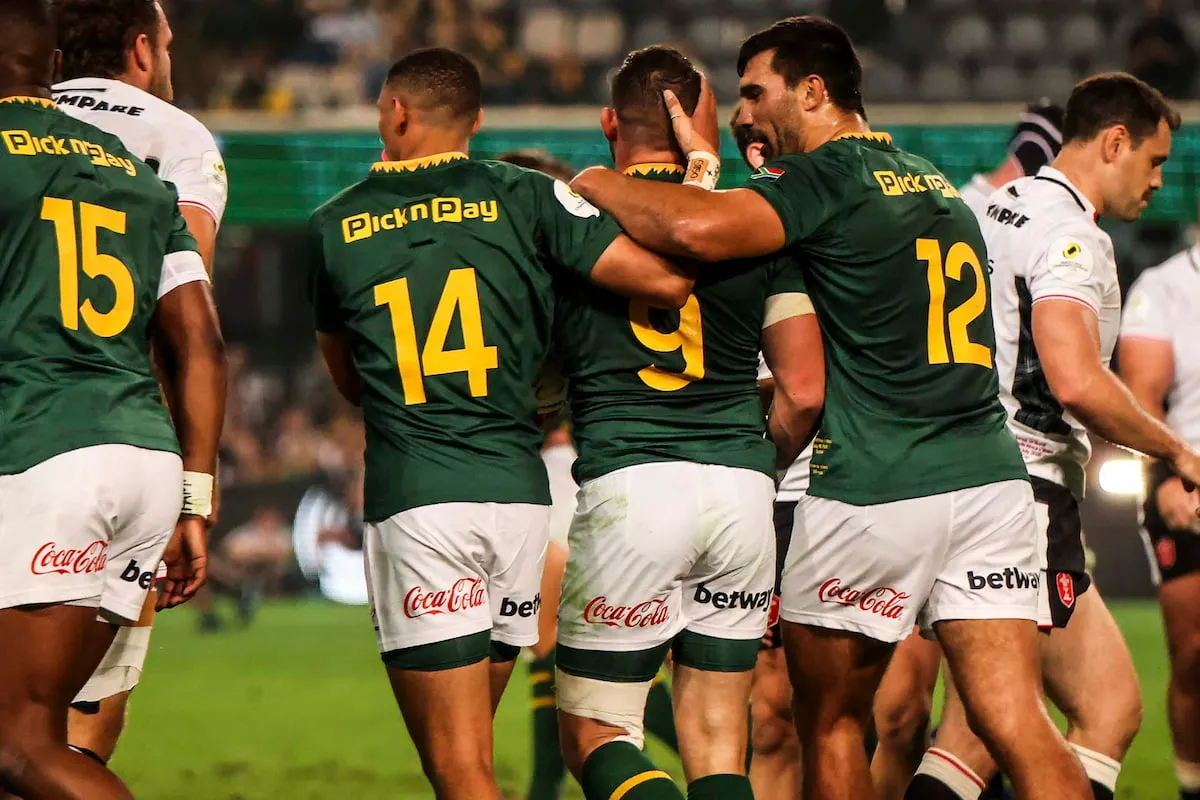 Rugby players in green jerseys with yellow numbers 14, 9, and 12 hug their teammates during a huddle on a stadium field, Coca-Cola and Betway logos visible on shorts/leathersays.