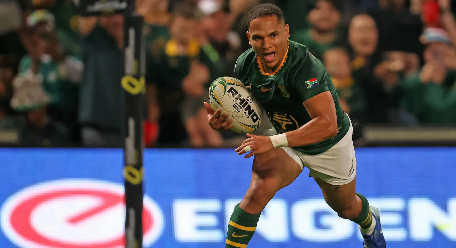 South African rugby player in green jersey runs with ball during a match, crowd in the stands behind him.