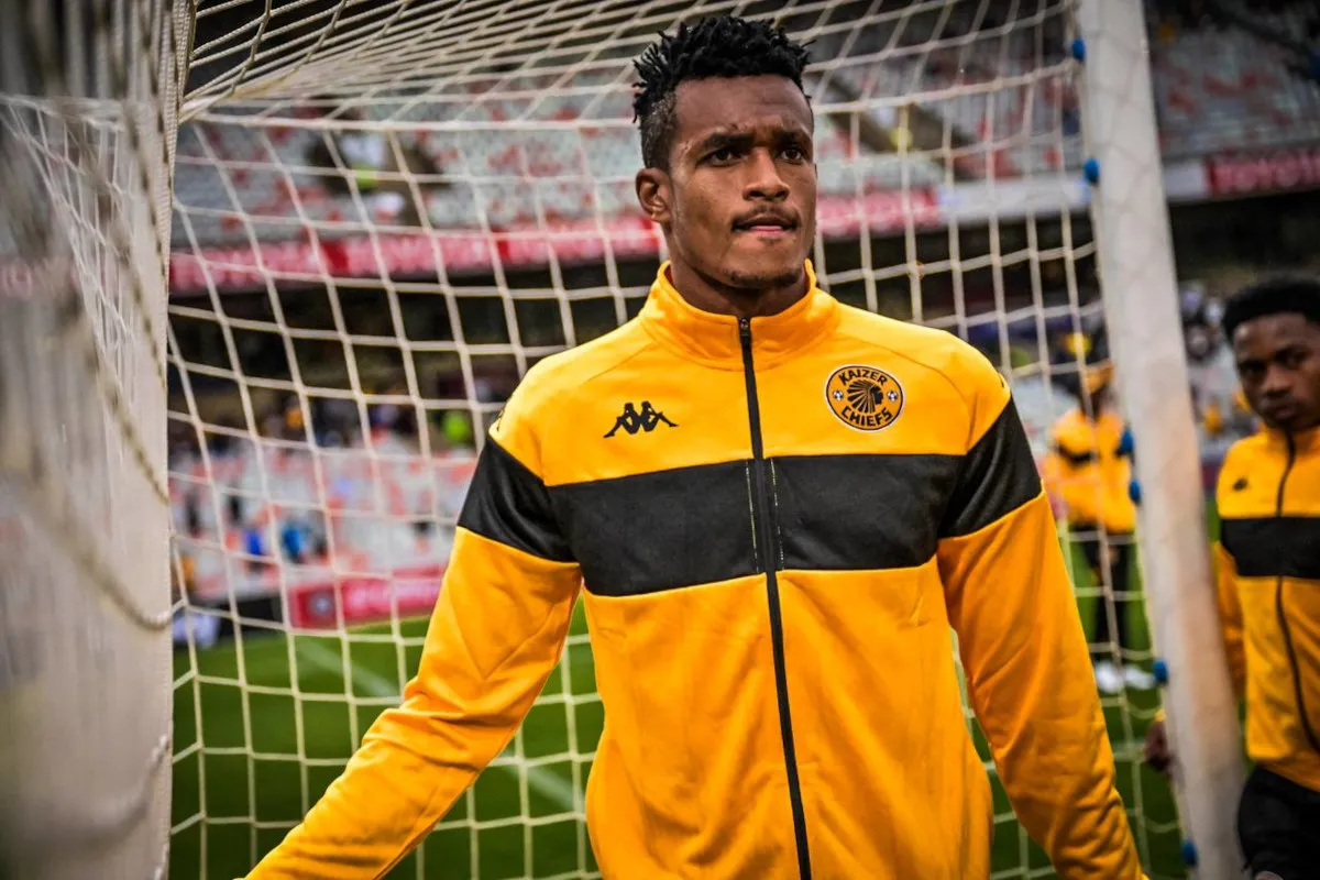 Footballer in a yellow and black Kaizer Chiefs warm-up jacket stands near the goal net on a football pitch, looking focused.