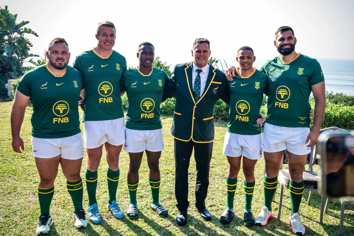 Six men in green rugby jerseys with yellow trim pose with a man in a dark blazer by the coast, smiling at the camera (team photo). (informative)
