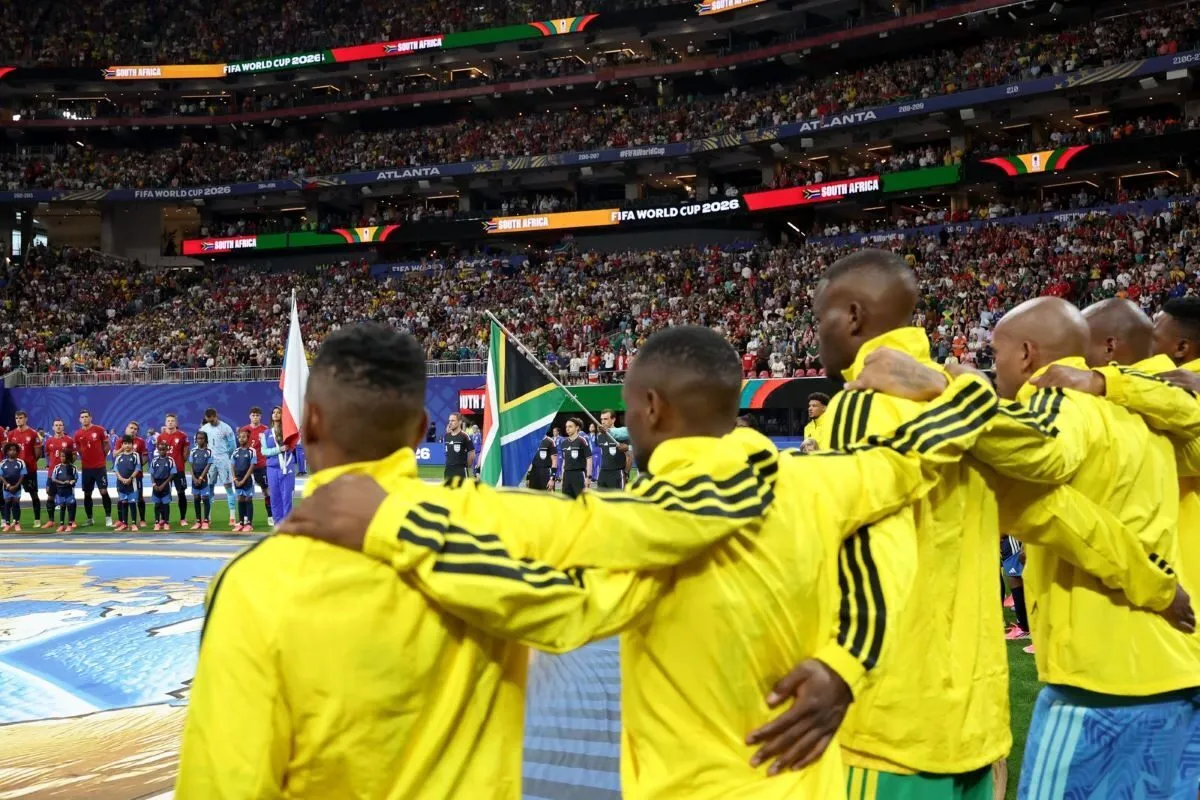 Football players in bright yellow jackets form a huddle on the pitch before a match, with a large stadium and flags in the background.