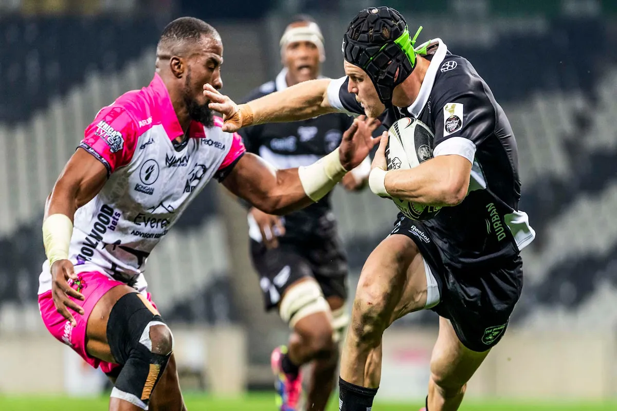 Rugby players clash mid-action: a black-kit player runs with the ball as a pink-and-white opponent reaches to tackle him.