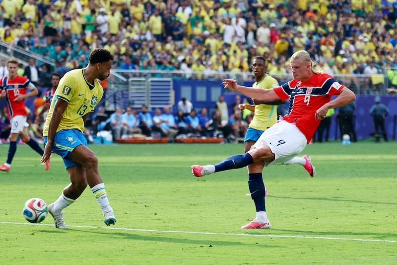 Brazilian player in yellow controls the ball as an opponent in red kicks during a soccer match with a crowded stadium in the background.