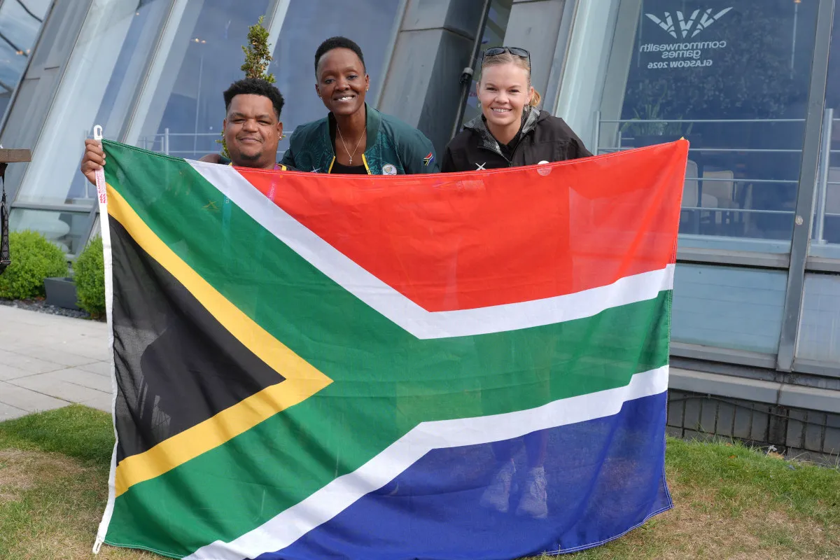 Three people smiling while holding a large South African flag in front of a glass building with windows.",