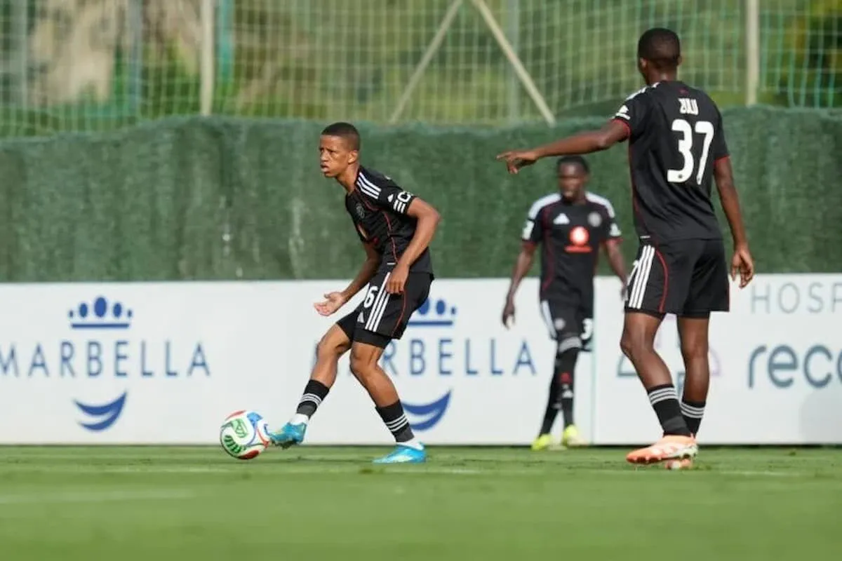 Soccer player in a black kit dribbles a ball on a green field as two teammates watch; advertising boards visible in the background.