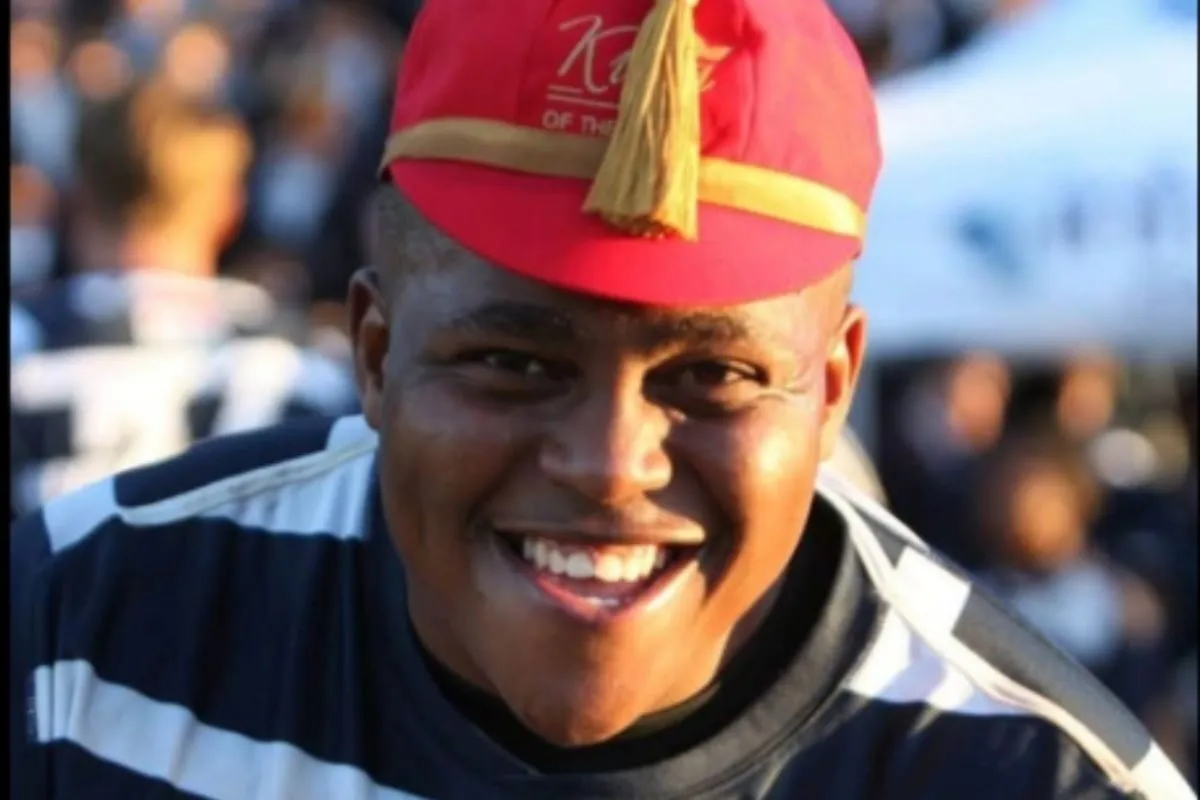 Close-up of a smiling man wearing a red hat with a yellow band and tassel at an outdoor event.
