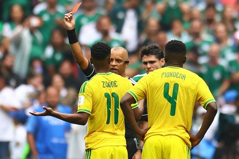 Soccer referee raises an orange card toward two players in yellow jerseys (numbers 11 and 4) as the crowd looks on from the stands behind them. Soccer referee raises an orange card toward two players in yellow jerseys (numbers 11 and 4) as the crowd looks on from the stands behind them.