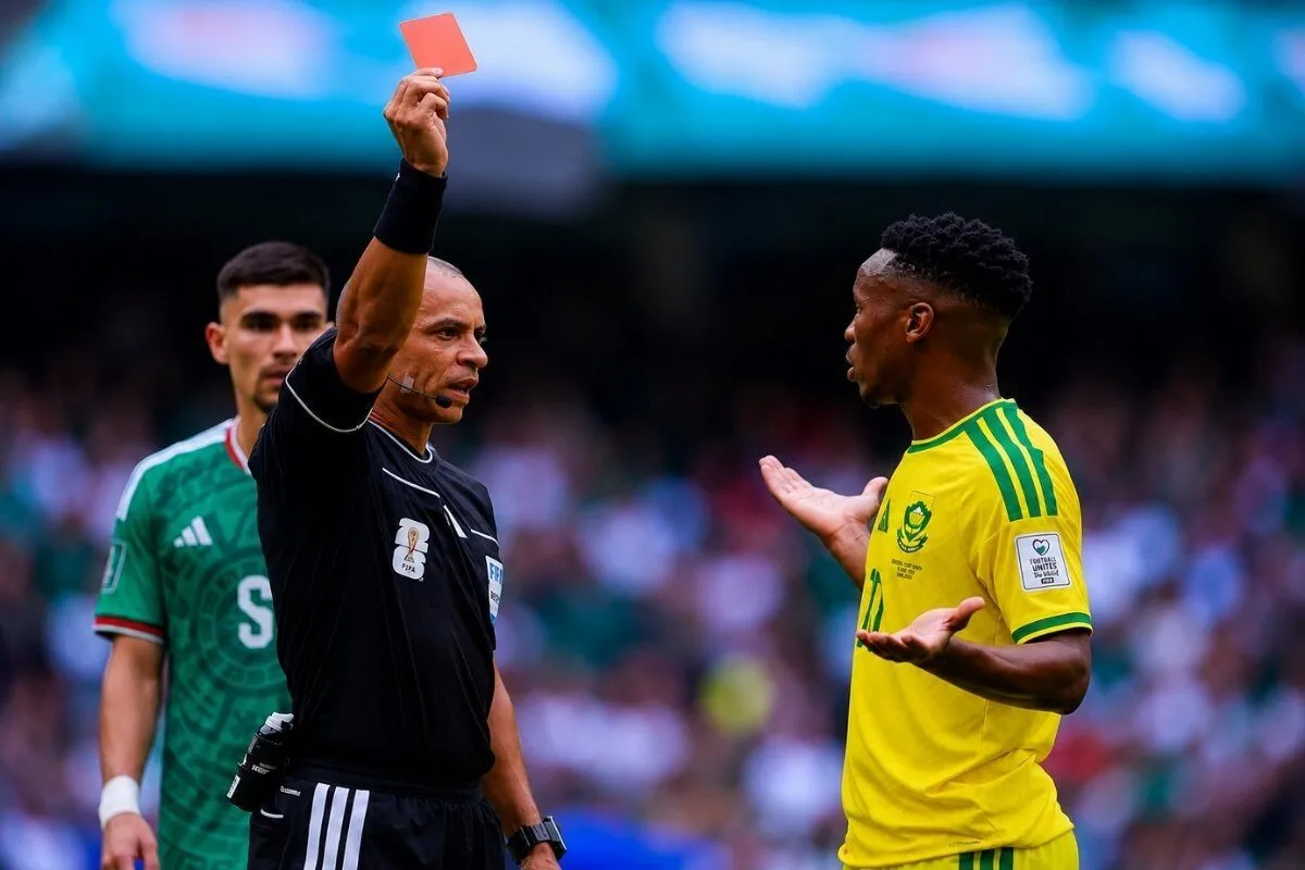 Soccer referee holds up an orange card toward a player in a yellow jersey during a match, while another green-jersey player watches. Soccer referee holds up an orange card toward a player in a yellow jersey during a match, while another green-jersey player watches.