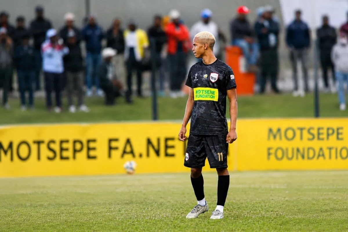 Soccer player with blond hair in a black kit standing on a grassy field, yellow sponsor boards and spectators in the background Soccer player with blond hair in a black kit standing on a grassy field, yellow sponsor boards and spectators in the background