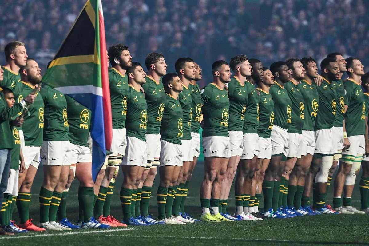 South Africa rugby team (Springboks) lined up on the field with a flag before a game, wearing green jerseys and white shorts.