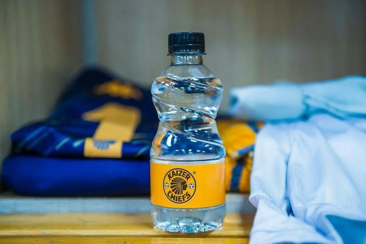 Water bottle with orange Kaizer Chiefs label on a wooden shelf, folded shirts in the background.