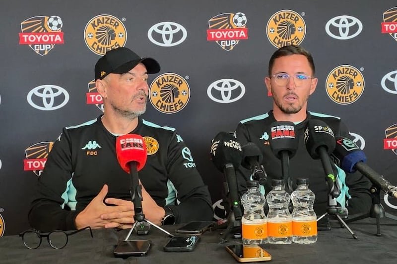 Two Kaizer Chiefs coaches sit at a press conference table with microphones, sponsor logos on the backdrop behind them.