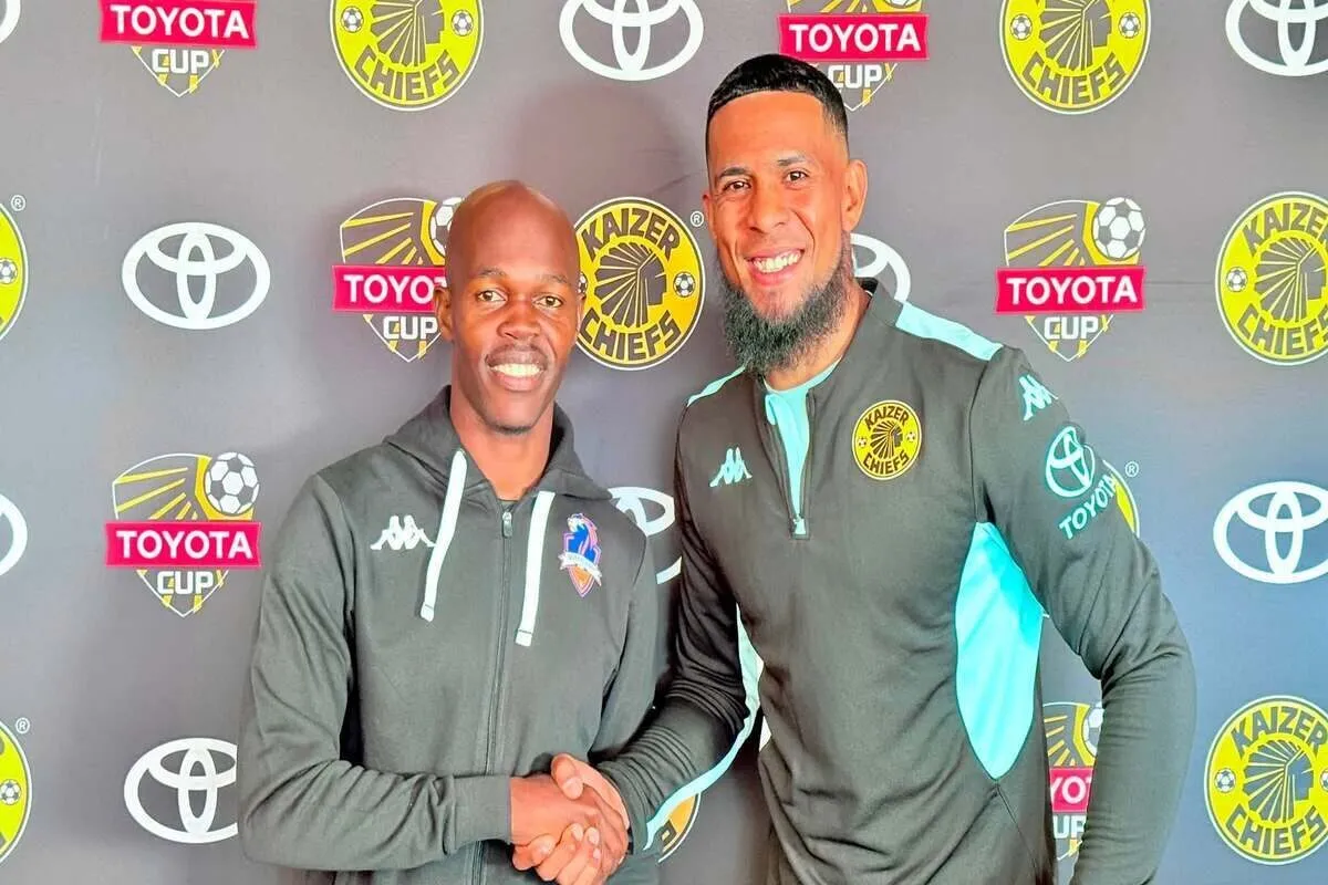 Two Kaizer Chiefs players in training tops shake hands and smile in front of a backdrop with Toyota Cup logos.