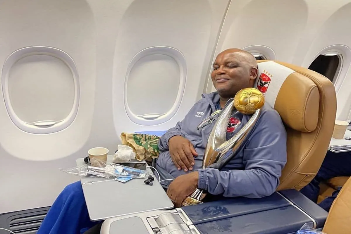 Man in a gray jacket sits in a plane seat, resting with a large gold soccer-ball trophy on his chest. Window seats visible behind him.