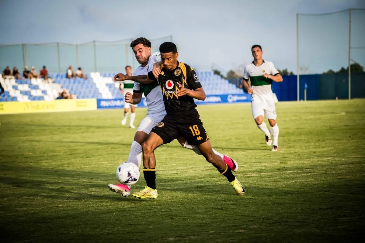 Two soccer players compete for the ball on a green field, one in black kit and the other in white, with a few spectators in the stands behind them.