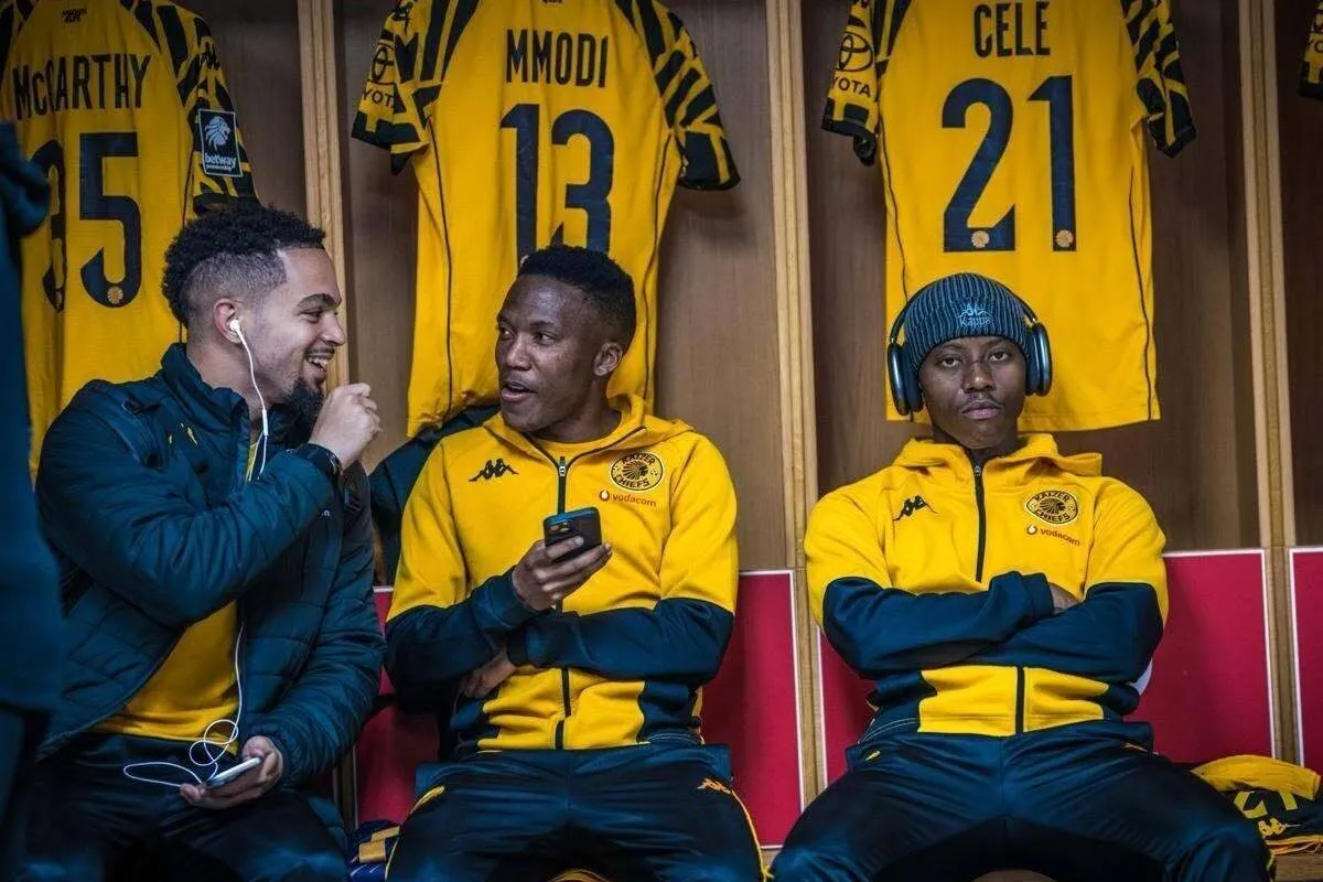 Three soccer players in yellow and black tracksuits sit in a locker room with hanging jerseys; two chat while the third wears headphones. Three soccer players in yellow and black tracksuits sit in a locker room with hanging jerseys; two chat while the third wears headphones.
