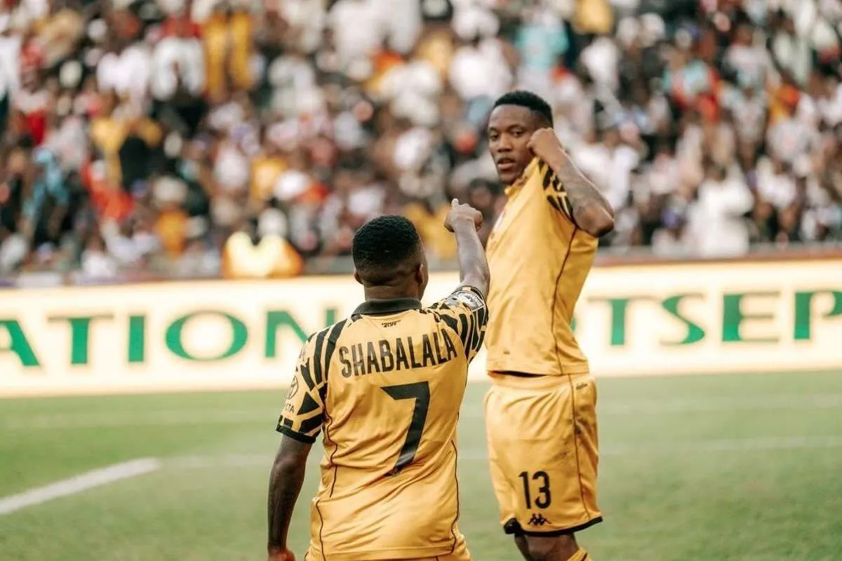 Two soccer players in gold jerseys celebrate on the field as a cheering crowd watches the action behind them, jersey #7 reads Shabalala. Two soccer players in gold jerseys celebrate on the field as a cheering crowd watches the action behind them, jersey #7 reads Shabalala.