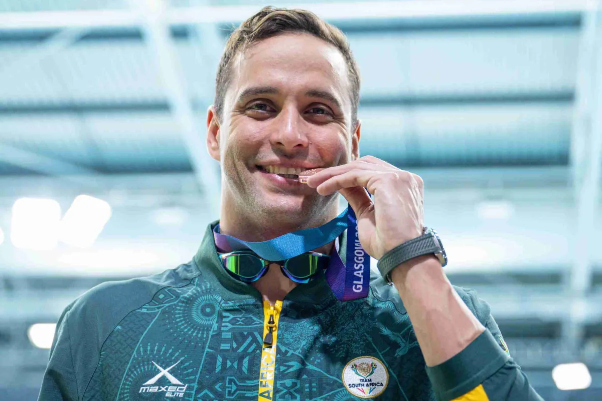 Smiling male athlete biting a medal, wearing a teal jacket with a Team South Africa badge and a Glasgow lanyard at a sports venue.
