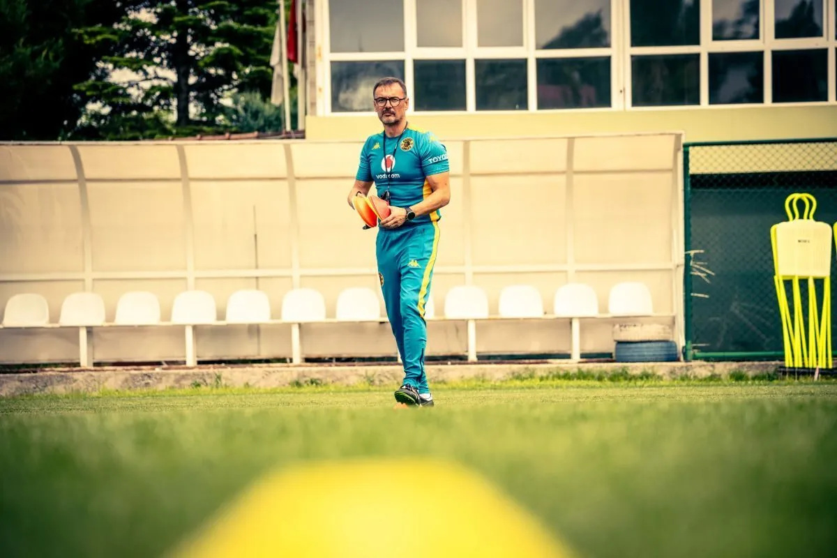 Athlete in turquoise training suit stands on grass holding an orange practice ball, with bench seating and a practice mannequin in background. Athlete in turquoise training suit stands on grass holding an orange practice ball, with bench seating and a practice mannequin in background.