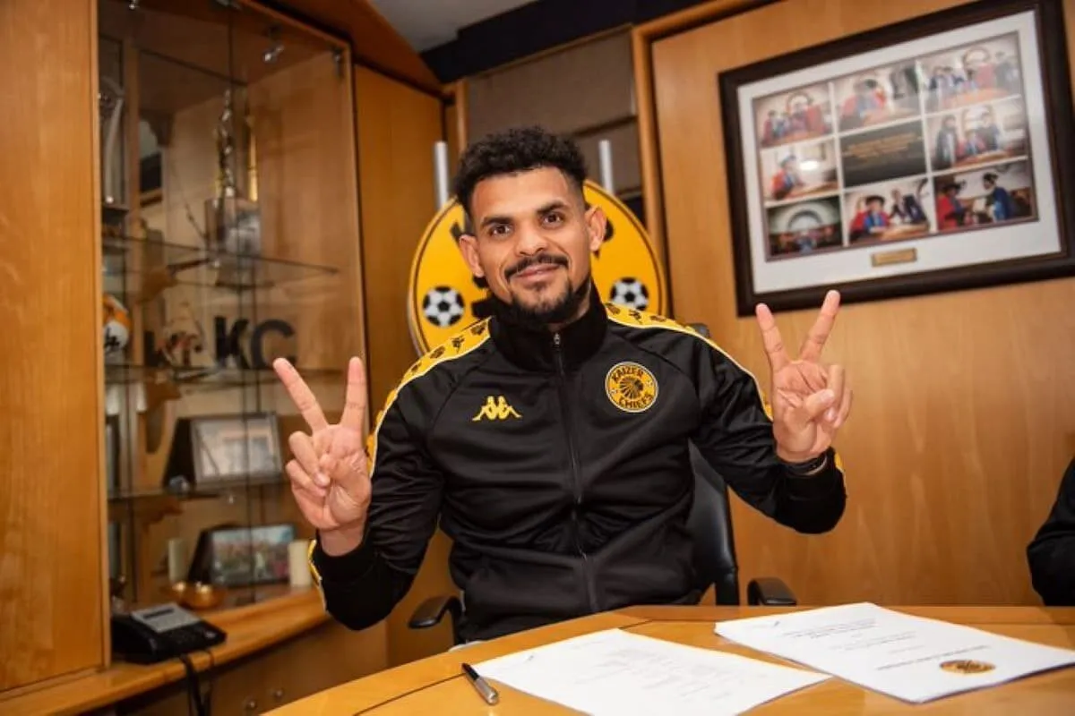 Man in a black and yellow sports jacket sitting at a desk, smiling and flashing peace signs with both hands in an office setting with a circular yellow club emblem behind him. Man in a black and yellow sports jacket sitting at a desk, smiling and flashing peace signs with both hands in an office setting with a circular yellow club emblem behind him.