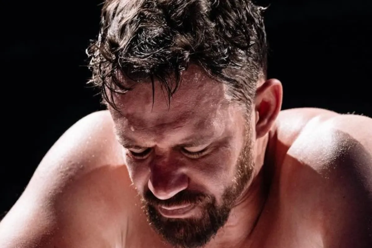 Close-up of a shirtless man with a beard looking downward, drenched in sweat with wet hair.