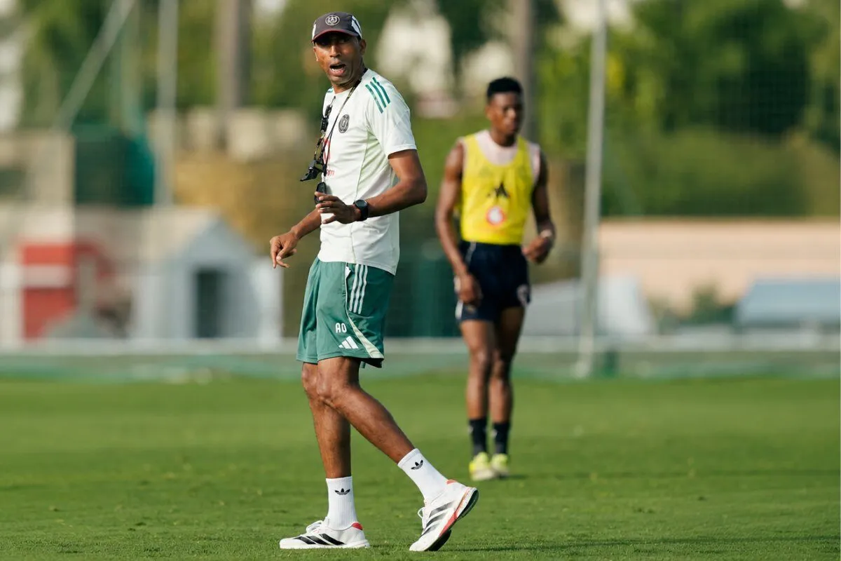 Soccer coach wearing a white shirt and green shorts on a training field, gesturing as a player in a yellow bib drills in the background.