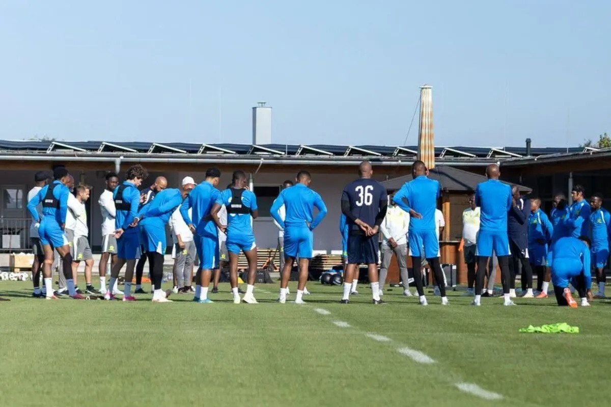 Soccer players in blue uniforms form a circle on a sunny training field, listening to coaches.