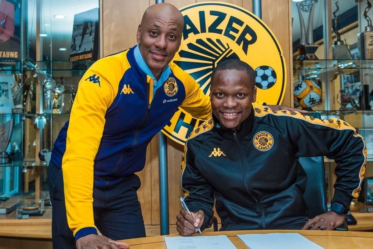 Two Kaizer Chiefs staff in yellow and blue jackets sign documents at a wooden desk with a trophy-filled backdrop behind them.