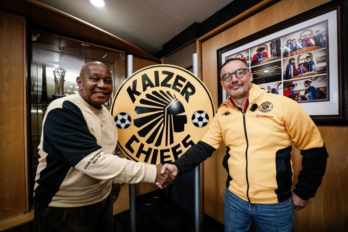 Two men shake hands in front of a Kaizer Chiefs emblem logo in a wood-paneled room, with framed photos on the wall behind them. Two men shake hands in front of a Kaizer Chiefs emblem logo in a wood-paneled room, with framed photos on the wall behind them.