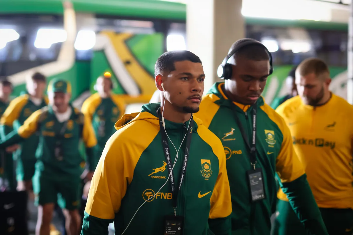 South African rugby players in green and gold jackets walk through a tunnel, wearing lanyards and earphones.