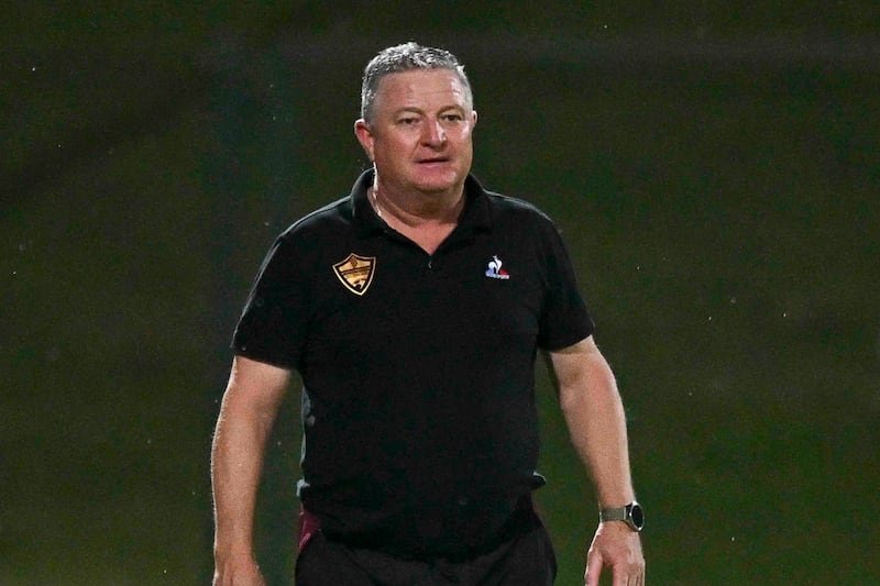 Man in a black polo on a sports field, wearing a watch, looking ahead as if coaching or supervising a game badge on shirt patch visible.