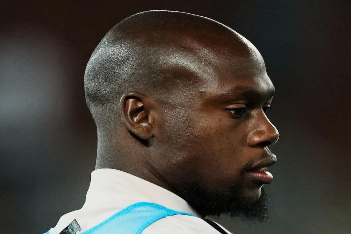 Side profile of a Black man with a trimmed beard wearing a white sports jersey with light blue trim.