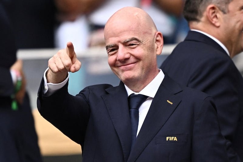 Smiling bald man in a dark suit points toward the camera at a formal event, wearing a FIFA badge on the chest.